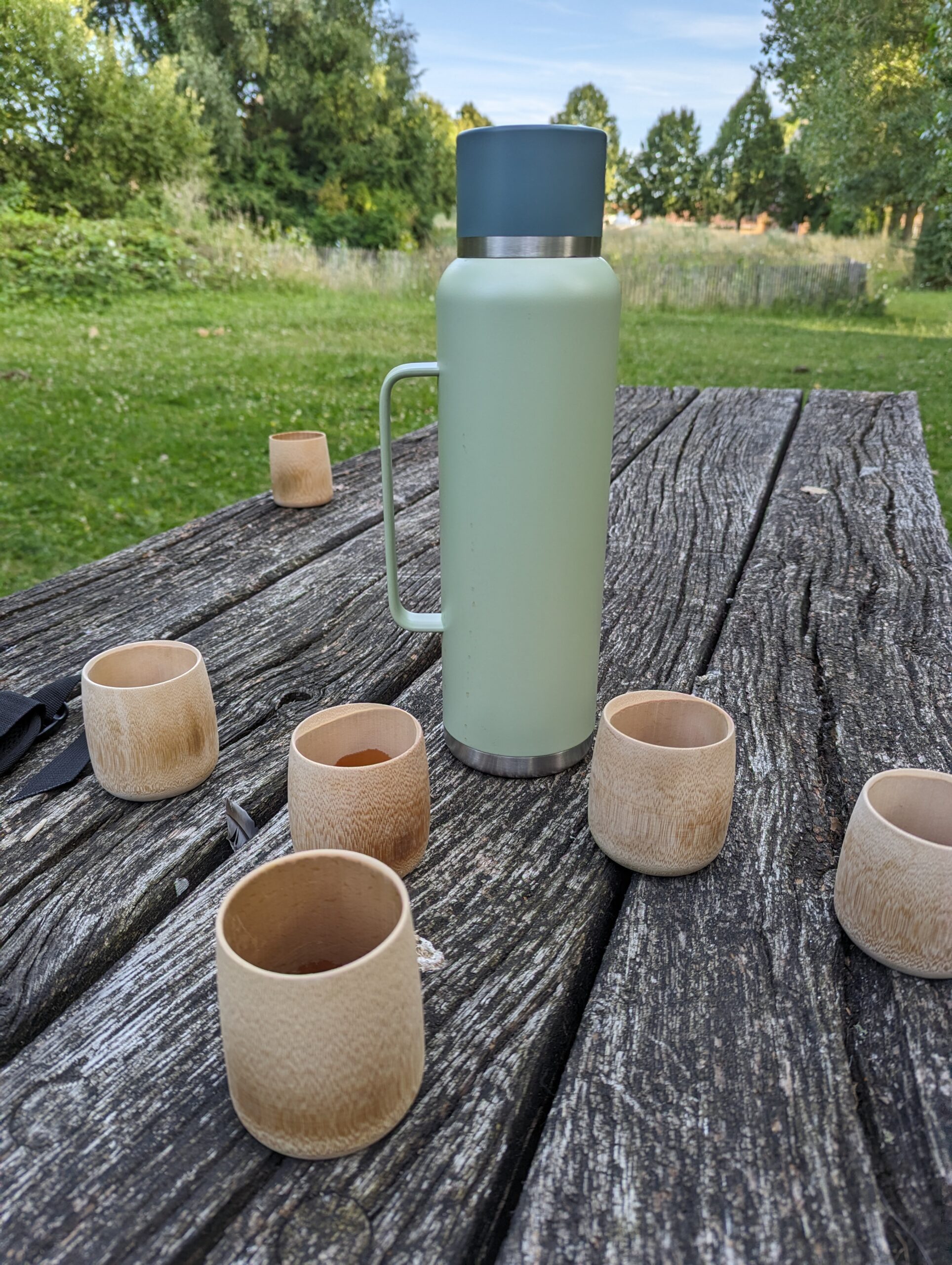 photo de gobelets en bambou et d'un thermos de thé, le tout sur une table en bois dans un parc, beaucoup de verdures